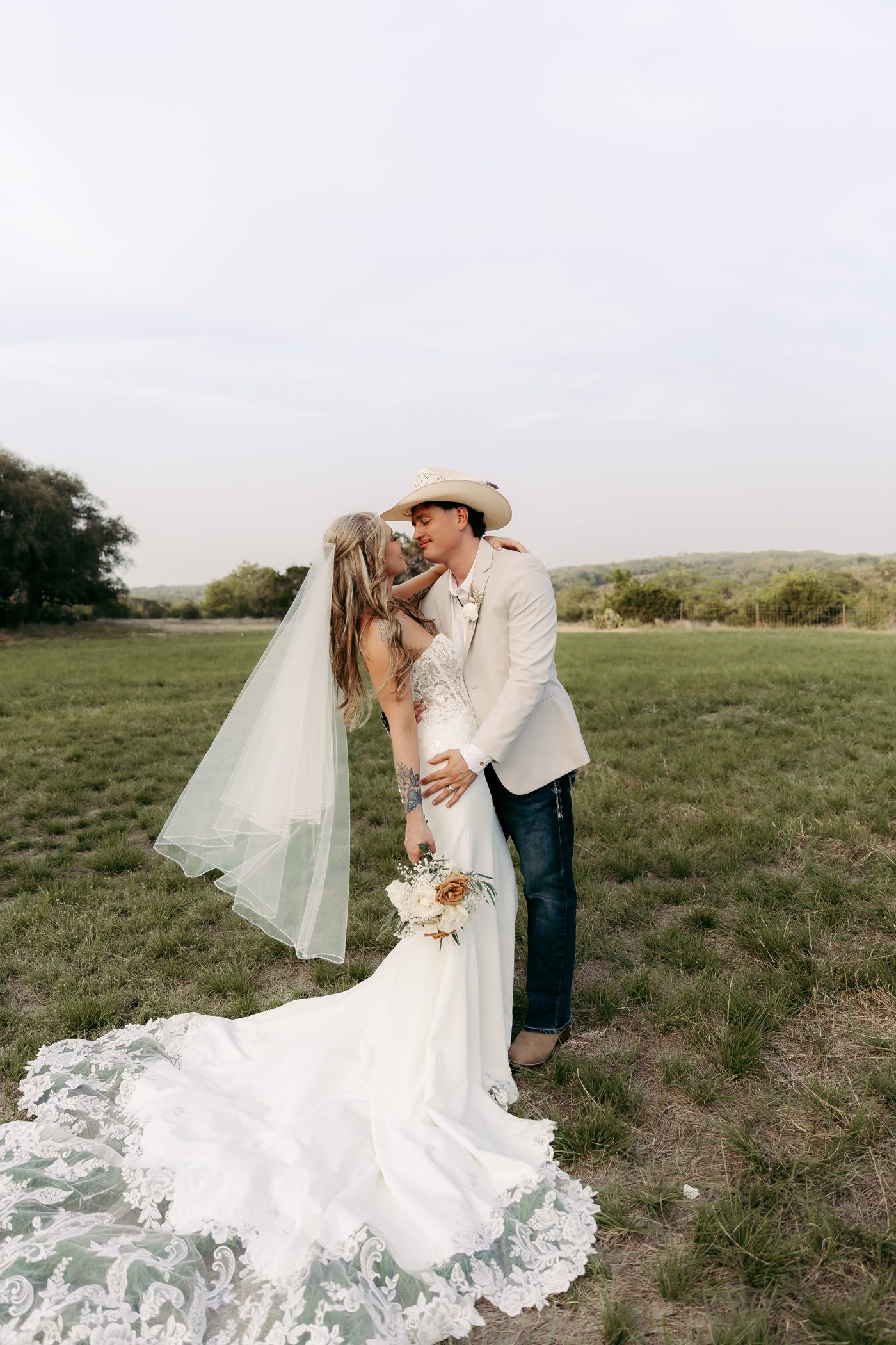 A bride and groom kissing in an open field at golden hour, her veil trailing behind