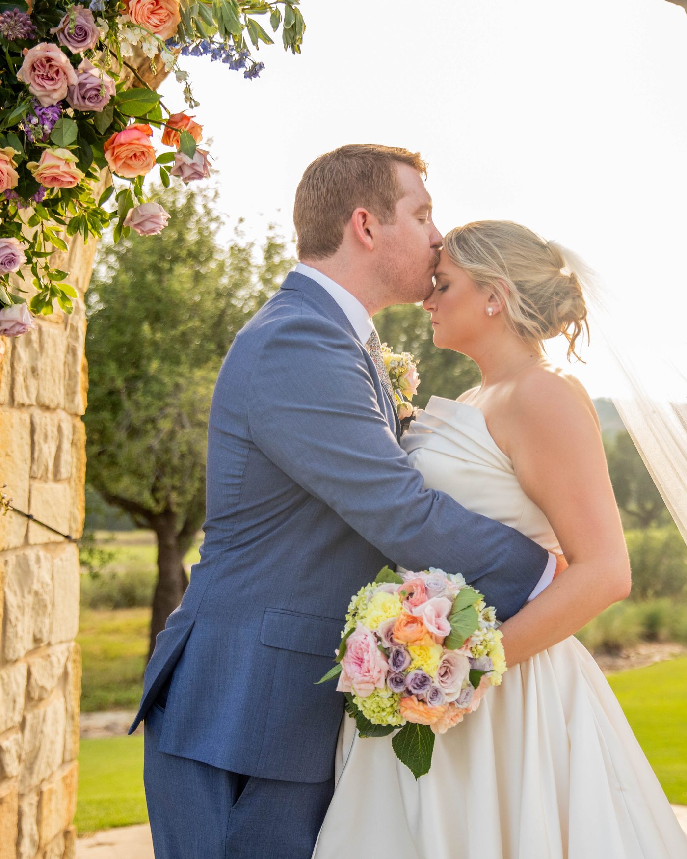 A groom kissing his bride beneath a flower-covered stone arch at golden hour